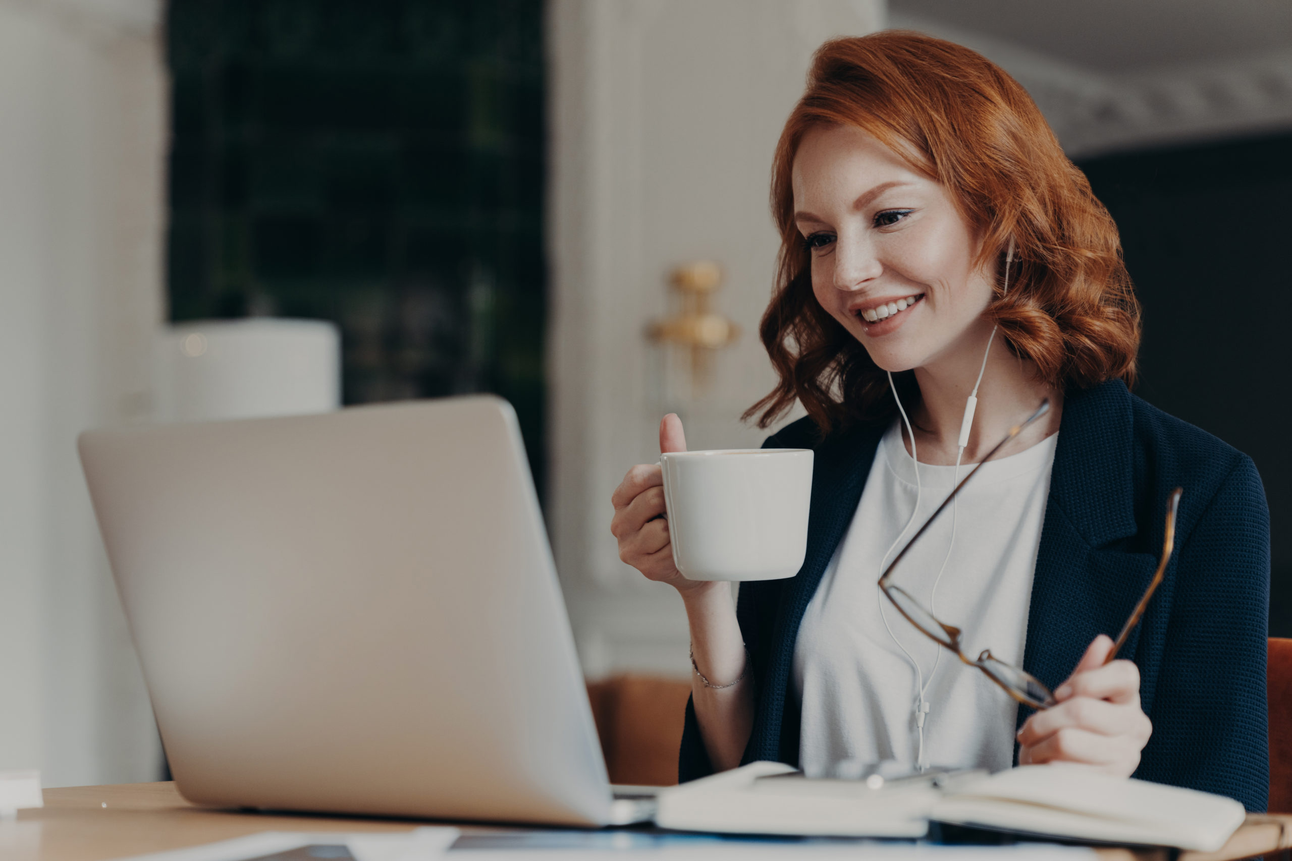 Intelligent female student has online course, concentrated in screen of laptop computer, learns foreign languages distantly, drinks tea or coffee, poses at desktop. Woman worker has video conference